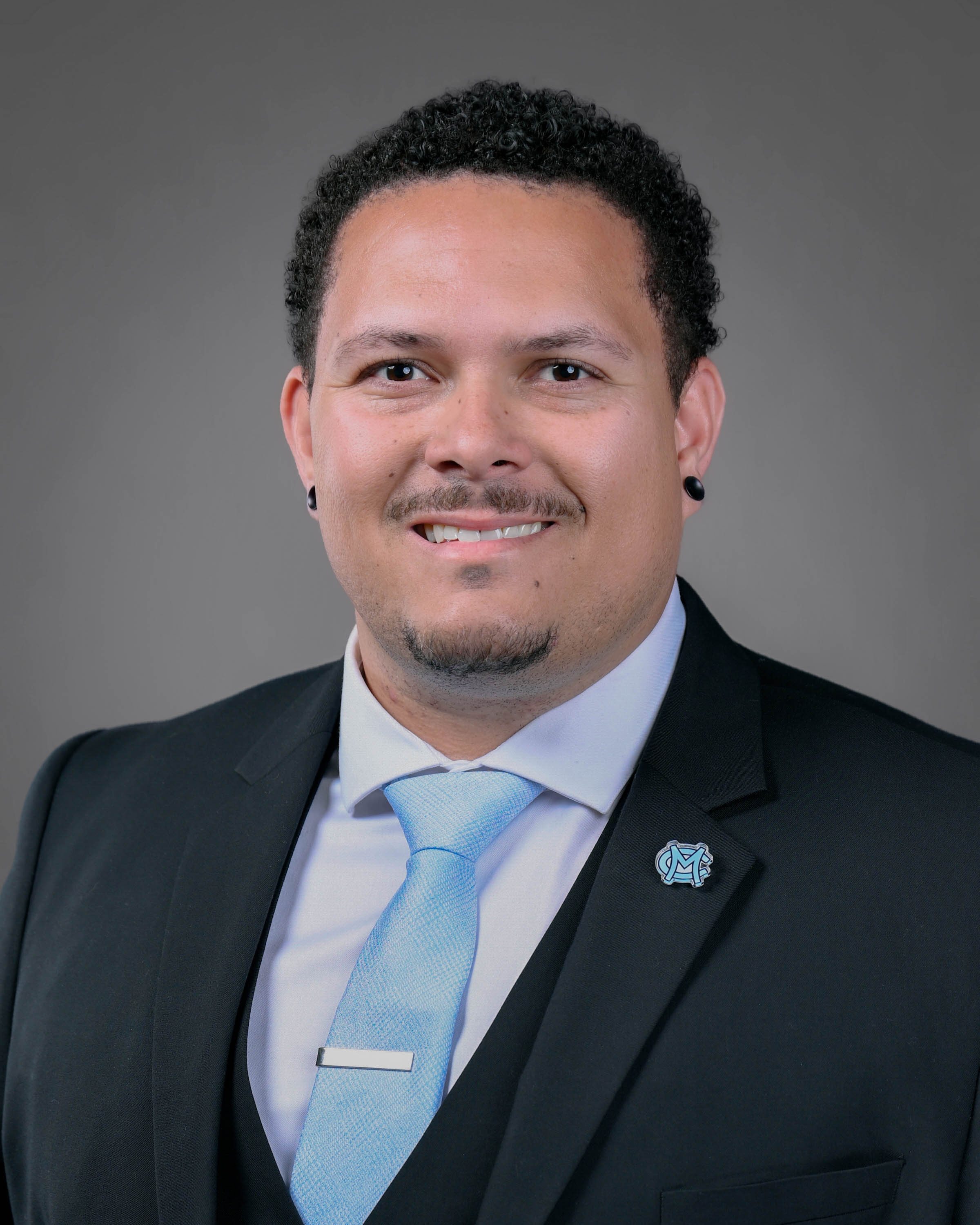 Smling man with short curly dark hair and a goatee in a dark suit wiht a light blue tie